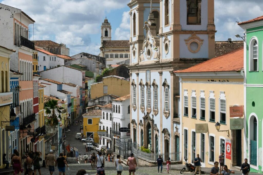 Vista de uma rua histórica com casarões coloniais coloridos, igreja de arquitetura barroca e pessoas caminhando pelo centro antigo de Salvador, Bahia. A cena representa o que é turismo cultural, destacando a valorização do patrimônio histórico, da arquitetura, da memória e das tradições locais. O ambiente preservado convida visitantes a conhecerem a história, a cultura e a identidade da cidade por meio de seus monumentos e espaços urbanos.