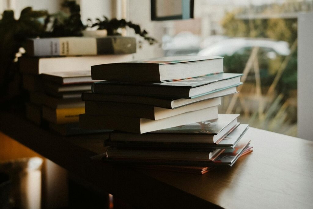 Pilha de livros sobre turismo cultural organizados sobre uma mesa próxima a uma janela, em um ambiente interno iluminado por luz natural. A fotografia destaca diferentes tamanhos e capas de livros empilhados, transmitindo uma atmosfera de leitura, estudo e pesquisa sobre turismo. Ao fundo, a cena aparece desfocada, com plantas e a paisagem externa visíveis através da janela.