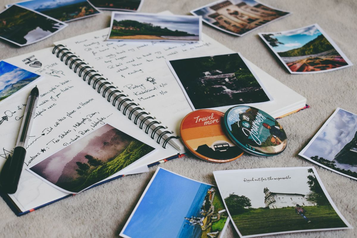 Um diário de viagem aberto sobre uma superfície de tecido claro, rodeado por fotos impressas de paisagens de viagem — montanhas, praias, cachoeiras e igrejas. As páginas do diário mostram anotações manuscritas com avaliações de destinos visitados, além de uma caneta apoiada ao lado. Dois bottons decorativos com frases sobre viagens completam a composição, transmitindo a sensação de nostalgia e planejamento de aventuras.